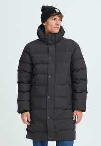 Svart pufferjakke med høy hals, knappelukking og lange ermer. Har en kviltet design og en avslappet passform, brukt over blå jeans.