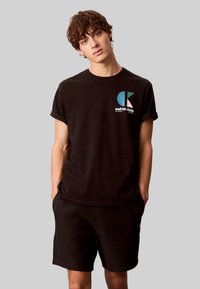 Schwarzes Baumwoll-T-Shirt mit umgekrempelten Ärmeln, versehen mit einem grafikdruck in Blau, Weiß und Türkis. Kombiniert mit schwarzen Shorts. Einfaches, lässiges Design.