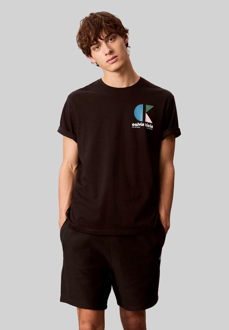 Schwarzes Baumwoll-T-Shirt mit umgekrempelten Ärmeln, versehen mit einem grafikdruck in Blau, Weiß und Türkis. Kombiniert mit schwarzen Shorts. Einfaches, lässiges Design.