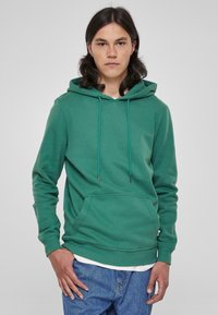 Felpa verde in morbido tessuto con tasca frontale, cappuccio regolabile con lacci, polsini a costine e orlo dritto. Indossata sopra una camicia bianca.