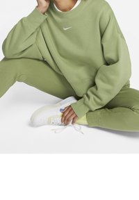 Olivgrön oversized sweatshirt med en vit Nike-logotyp, matchad med byxor i samma stil och vita sneakers med pastellaccenter.