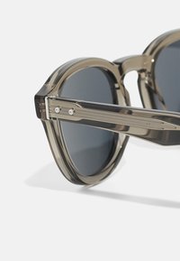 Grau getönte Sonnenbrille mit einem halbtransparenten, kantigen Kunststoffrahmen und metallischen Akzenten an den Bügeln, die ein strukturiertes Design aufweisen.