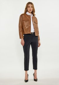 Braune Leder-Jacke in cropped Länge mit zwei Vorderseiten-Taschen, Knopfverschluss und glatter Textur, kombiniert mit schwarzen maßgeschneiderten Hosen und schwarzen Stöckelschuhen.