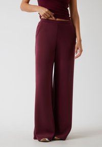 Vrouw gekleed in burgundy hoge taille, wijde broek met bijpassend mouwloos topje, staande op een effen lichtgrijze achtergrond.