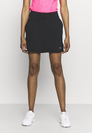 Kvinde iført et sort Nike atletisk skørt, lyserød ærmeløs top og hvide sportssko, der står mod en ensfarvet hvid baggrund.