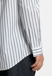 Weißes Langarmhemd mit schmalen blauen vertikalen Streifen, geknöpftem Ärmelbündchen und geschwungenem Saum; der Stoff wirkt glatt und leicht.