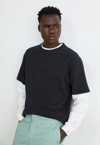 Short-sleeve black t-shirt with a crew neckline and subtle embroidered detail, layered over a white long-sleeve shirt, paired with mint green pants.