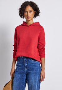 Femme avec des cheveux courts et bouclés portant un sweatshirt rouge et un jean bleu, tenant un sac à main beige devant un fond clair.