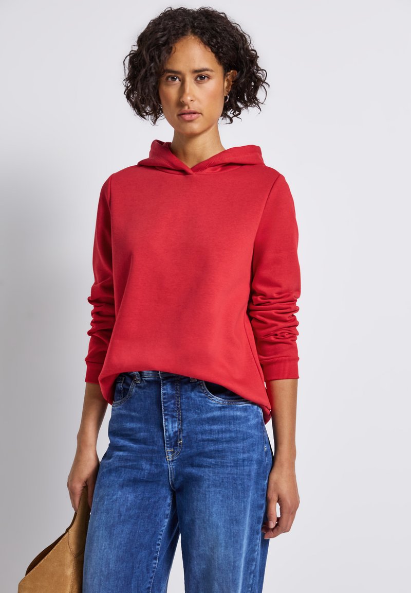 Femme avec des cheveux courts et bouclés portant un sweatshirt rouge et un jean bleu, tenant un sac à main beige devant un fond clair.