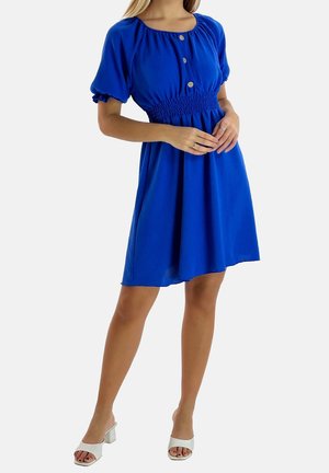 Femme portant une robe bleu roi longueur genou avec des manches bouffantes, boutonnée devant, taille smockée, et des sandales blanches à talons, debout les mains jointes.