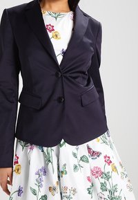 Námornický blazer s přiléhavým designem, špičatými klopami, na dvě knoflíky a předními kapsami. Nesený přes bílé šaty s barevnými květinovými vzory.