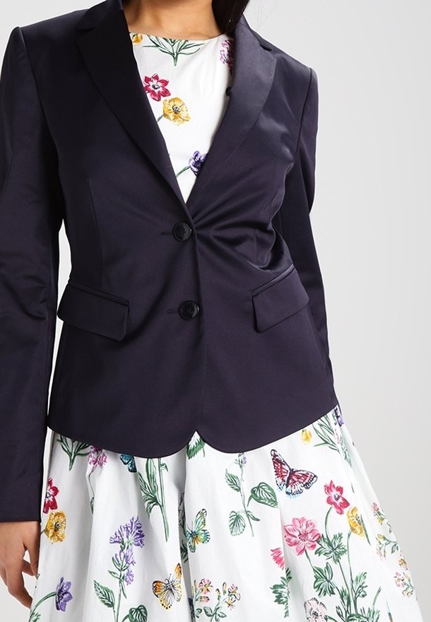 Námornický blazer s přiléhavým designem, špičatými klopami, na dvě knoflíky a předními kapsami. Nesený přes bílé šaty s barevnými květinovými vzory.