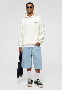 Witte hoodie met een voorzak en geborduurde tekst, gecombineerd met lichtblauwe denim shorts, zwarte sneakers en een zonnebril, terwijl er een tote bag wordt vastgehouden.