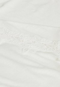 Bílá látka s jemnou texturou má na jedné straně delikátní vyšívanou krajku. Krajka zobrazuje květinový vzor.