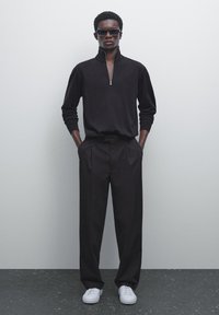 Zwarte ritsvest met een hoge kraag, gecombineerd met losse zwarte broek en witte sneakers, tegen een neutrale achtergrond.