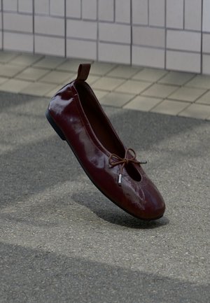 Särav tumepunane baleriiniking, millel on peal väike lips, seisab püsti tekstuurilisel pinnal lähestikku plaatidest seina taustal.