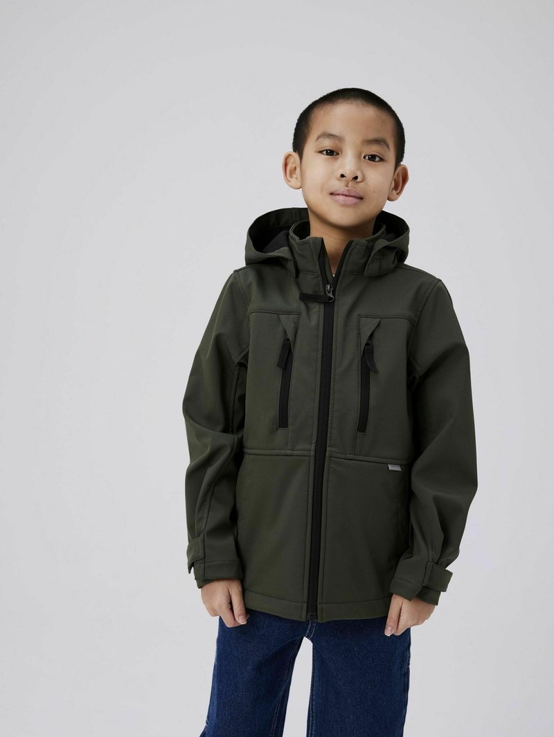 Veste imperméable vert foncé avec une capuche, dotée d'une fermeture éclair à l'avant et de deux poches poitrine avec des accents noirs, associée à un jean bleu.