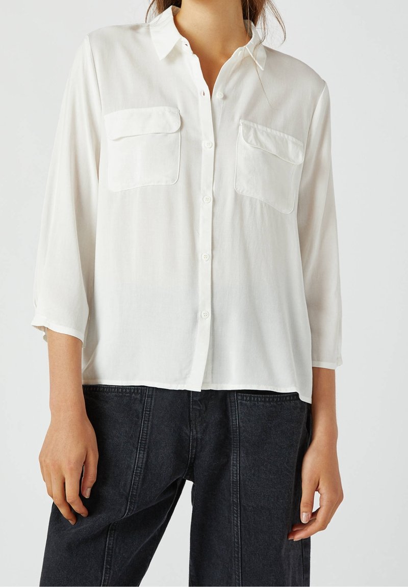 Biała koszula z długim rękawem uszyta z lekkiego materiału, z dwiema kieszeniami na piersi. Noszona z ciemnymi dżinsami.