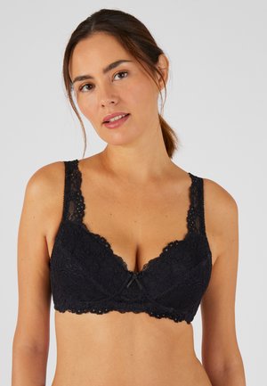 Femme aux cheveux bruns attachés, portant un soutien-gorge en dentelle noire, faisant face avec une expression neutre sur un fond uni.