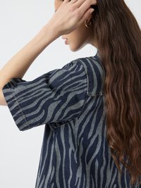 Camicia di jeans con un motivo a strisce zebra blu e grigie, maniche corte e dettagli di cucitura visibili. La texture appare liscia e informale.