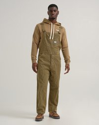Salopette en velours côtelé vert olive avec bretelles réglables, poche sur la poitrine et coupe décontractée, portée sur un hoodie marron et des baskets marron.