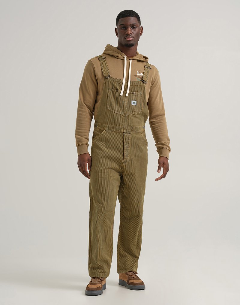 Salopette en velours côtelé vert olive avec bretelles réglables, poche sur la poitrine et coupe décontractée, portée sur un hoodie marron et des baskets marron.