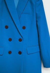 Manteau bleu croisé avec revers crantés, deux poches avant et cinq boutons ronds marron foncé, présentant une texture de tissu lisse.