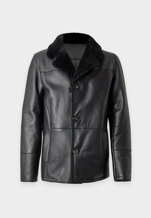 Homme portant une veste en cuir noire avec col en fourrure, un pull marron, un pantalon en cuir noir, une ceinture noire et des bottines noires, debout sur un fond uni.