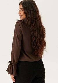 Blusa marrone a maniche lunghe con una texture liscia, caratterizzata da polsini svasati e una scollatura modesta, abbinata a pantaloni neri.