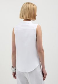 Donna con capelli biondi corti che indossa una blusa bianca senza maniche, pantaloni bianchi e un grande braccialetto d'argento, in piedi davanti a uno sfondo neutro.