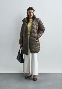 Καφέ γεμάτο παλτό με γυαλιστερή επιφάνεια, που διαθέτει υψηλό γιακά και γεωμετρικά σχέδια. Συμπληρώνεται με κίτρινη μπλούζα και λευκά παντελόνια φαρδιάς γραμμής.