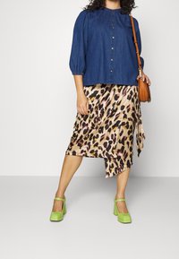 Donna che indossa una camicia blu con bottoni, una gonna in satin con stampa leopardata, tacchi verde lime e porta una borsa a tracolla marrone.