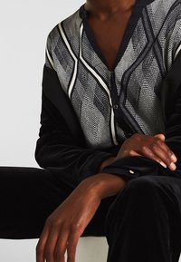 Persona che indossa una blusa a motivi bianco e nero con maniche in velluto nero, seduta con le braccia incrociate e una postura rilassata.