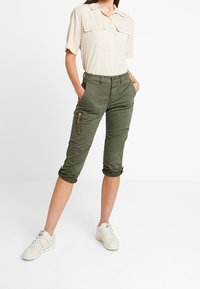Světle béžová košile s krátkým rukávem a dvěma náprsními kapsami; olivově zelené cropped cargo kalhoty s kapsou na zip; bílé tenisky.