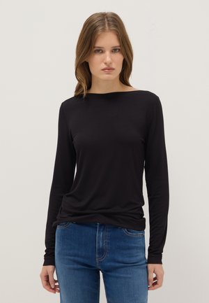 Young woman with wavy light brown hair wearing a black long-sleeve top and blue jeans, standing against a plain light background.