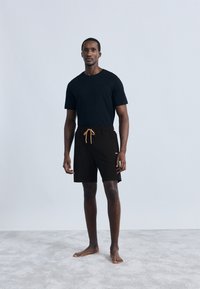 Μαύρο μπλουζάκι και μαύρο σορτς με κορδόνι και ανοιχτό καφέ λεπτομέρεια. Το μοντέλο στέκεται ξυπόλυτο σε ανοιχτό γκρι χαλί, κοιτάζοντας μπροστά.