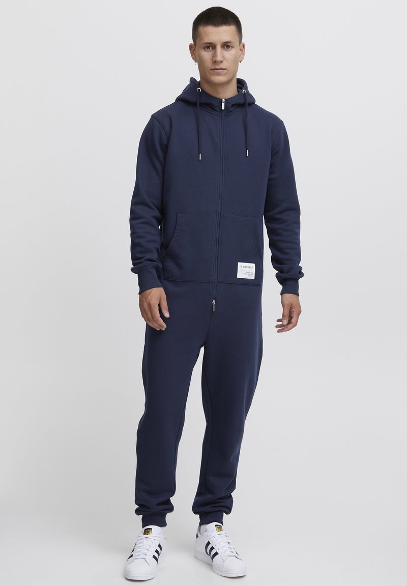 Conjunto de sudadera con cremallera y joggers en azul marino, hecho de una tela suave. Presenta un bolsillo tipo canguro, cordones y un pequeño parche con un logo.