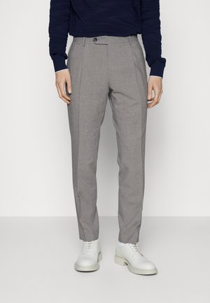 Mężczyzna ubrany w szare, szyte na miarę spodnie, granatową bluzkę z długim rękawem i białe eleganckie buty, stojący na jednolitym jasnym tle.