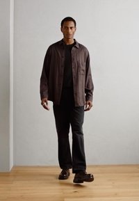 Camicia marrone con colletto in un tessuto morbido e texturizzato, abbinata a una camicia nera e jeans scuri. Scarpe eleganti completano l'outfit. Sfondo minimalista.