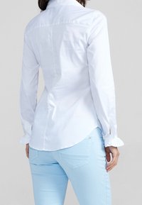 Weißes Hemd mit langen Ärmeln und Rüschenmanschetten, präsentiert mit hellblauen Hosen, die eine glatte Textur und eine taillierte Passform hervorheben.