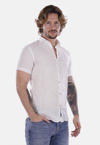 Camicia bianca di lino a maniche corte con placket abbottonato e una texture morbida. Presenta una vestibilità rilassata e bottoni chiari a contrasto.