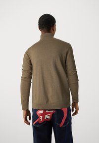 Brązowy sweter z dzianiny z wysokim kołnierzem, długimi rękawami i ściągaczem u dołu. Połączony z ciemnymi dżinsami z czerwonym nadrukiem graficznym.