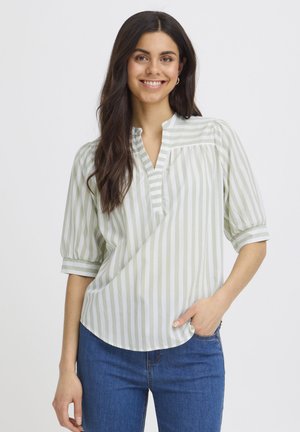 Usmievavá žena s dlhými tmavými vlasmi, oblečená v svetlozelenej blúzke s bielymi pruhmi a modrých džínsoch, s rukou v vrecku, na bielom pozadí.