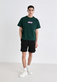 Grønn bomulls t-skjorte med rosa grafikk, kombinert med sorte shorts. Hvite sneakers og ankelsokker fullfører antrekket. Enkelt design, uformell passform.