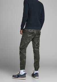 Cargo kalhoty ve tmavě zeleném kamuflážovém vzoru s elastickými manžetami, v kombinaci se šedými teniskami. Námořnický svetr má texturovaný pletený design.