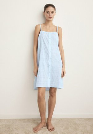 Jeune femme debout pieds nus contre un mur pâle, portant une robe bleu clair sans manches avec des bretelles fines et des boutons à l'avant.