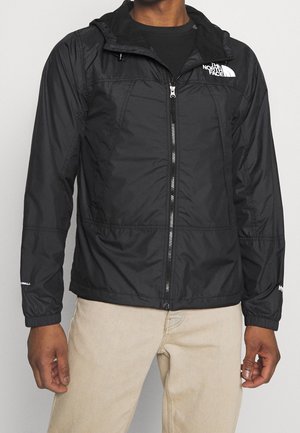 Mann trägt eine schwarze North Face Windjacke mit Reißverschluss und Kapuze sowie beige Hose vor neutralem Hintergrund.