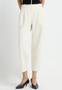 Crème-kleurige high-waisted broeken met plooien, taps toelopende benen en zijzakken, gecombineerd met zwarte lage hakken. Soepele textuur.