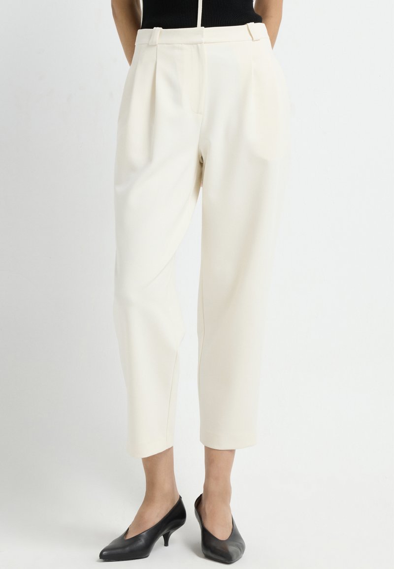 Crème-kleurige high-waisted broeken met plooien, taps toelopende benen en zijzakken, gecombineerd met zwarte lage hakken. Soepele textuur.