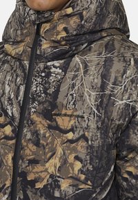 Kurtka puchowa w kamuflażu z brązowymi, szarymi i oliwkowymi kolorami; posiada wysoki kołnierz, zamek błyskawiczny i detal z logo na klatce piersiowej.